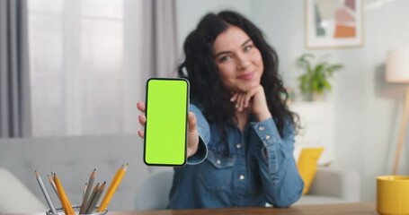 Beautiful young woman looking at camera and showing chroma key indoors. Pretty Caucasian female holding smartphone with green screen and smiling at home. Vertical position. Technology. - Powered by Adobe