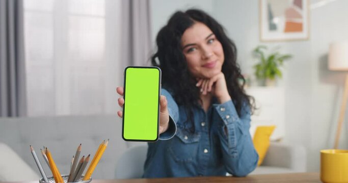 Beautiful young woman looking at camera and showing chroma key indoors. Pretty Caucasian female holding smartphone with green screen and smiling at home. Vertical position. Technology.
