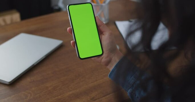 Back view of curly haired Caucasian lady sitting at table and video calling friend. Young woman holding smarphone with green screen, chroma key in home office. Technology, social media, facetime app.
