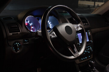 The driver's seat in the cabin of a modern car with a leather steering wheel and expensive equipment. Night photography with interior lighting