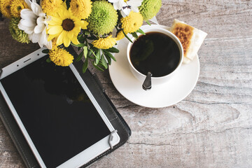 a Cup of coffee with the tablet turned off next to a bouquet of flowers on a wooden table.