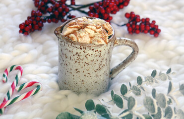 Hot chocolate in a mug topped with marshmallows and whipped cream on white knitted surface. Red berries branch, candy canes and eucalyptus branch around the mug. 