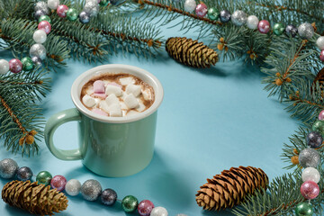 A mug with hot chocolate and marshmallows on a blue background, decorated with fir branches, garlands and beads.