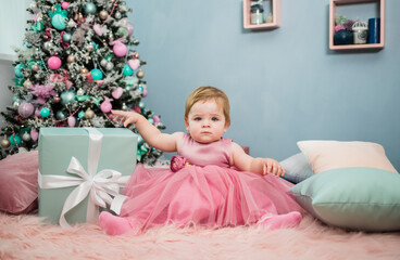 a little girl in a festive pink dress sits on a fur blanket with a large blue gift on the background of a Christmas tree