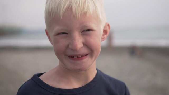Close-up of the face of a beautiful blonde boy against the background of the washed-out sea and beach.The camera slowly approaches the cheerful face of the child