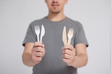 zero waste and eco friendly concept - man comparing plastic and wooden forks and knives over grey...