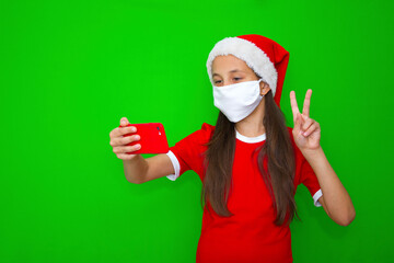 Fototapeta na wymiar A girl with dark hair in a Santa hat in a medical mask, in a New Year's outfit with a mobile phone. Shows victory. Isolated green background.