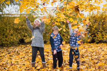 Three children are playing with yellow fallen leaves. Siblings are throwing maple leaves up in autumn park. Autumn and fall concept.