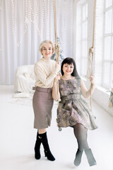 Pretty Caucasian brunette woman, sitting on swing in decorated room with Christmas tree and big window and enjoying time with her sister or friend, standing nearby. Concept of New Year, Christmas