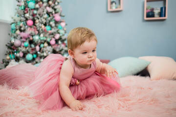 Fototapeta premium a playful little girl in a festive pink dress crawls on a furry pink blanket on the background of a Christmas tree