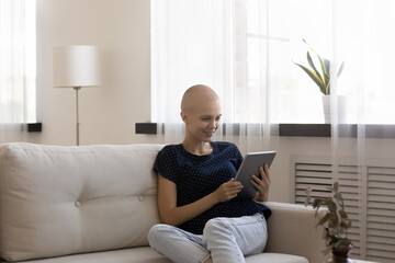 Happy hairless woman holding computer tablet, sitting on couch, enjoying leisure time at home,...