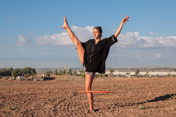 Female flow artist dancing with a Hula hoop outdoors, with one leg on the ground. Wide shot in...