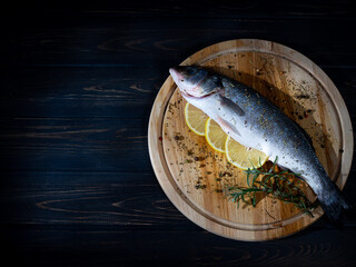 A whole fresh fish with slices of lemon, rosemary and spices on a round cutting board. Dark wooden...