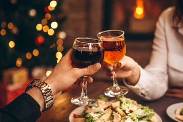 hands with glasses clinking against the background of Christmas tree lights and bonfires from a home fireplace over a table with delicious dishes