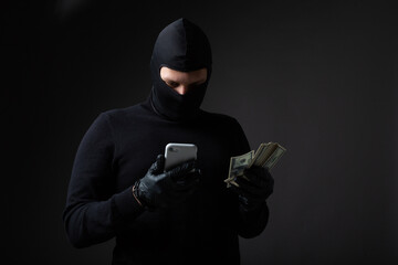 robber in black mask with money and phone in hands on black background