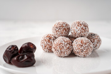 Candy balls made of dates and coconut shavings in a white dish on a light background near the dates