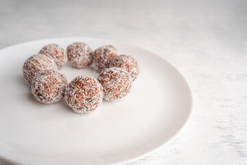 A white plate with round sweets made of dates and coconut shavings on a light wooden background
