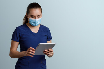 Mujer latina, con uniforme de enfermera usando cubrebocas y careta, sosteniendo una tableta digital en las manos, y un fondo color azul con espacio para texto