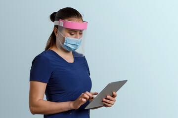 Mujer latina, con uniforme de enfermera usando cubrebocas y careta, sosteniendo una tableta digital en las manos, y un fondo color azul con espacio para texto © Edgar1 BJ