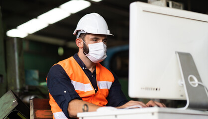 Young caucasian engineering man worker working on digital tablet computer at manufacturing. Worker man wearing face mask prevent covid-19 virus and protective hard hat.