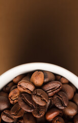 Coffee beans, with ground (selective focus on coffee beans) and white ceramic cup, brown background.