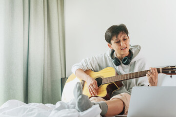 Lady with is singing song and playing on acoustic guitar with laptop on bed in apartment.