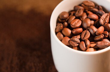 Coffee beans, with ground (selective focus on coffee beans) and white ceramic cup, brown background.