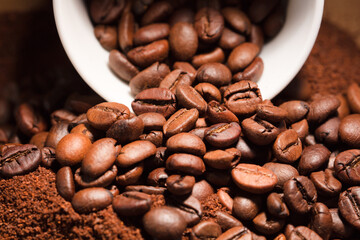 Coffee beans, with ground (selective focus on coffee beans) and white ceramic cup, brown background.