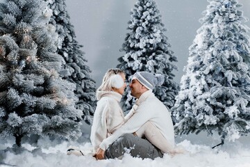 Fototapeta na wymiar Man kiss and hug his woman and snow falls. New year love story. People weared wearing fur headphones, hats, white sweaters.