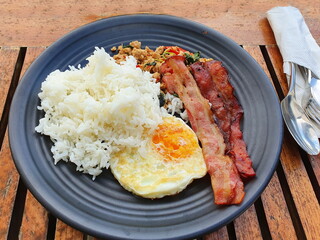 Breakfast in black ceramic plates, consisting of steamed rice, bacon, fried egg, stir-fried minced pork with basil, served on a wooden table with a stainless steel spoon and fork in a white tissue bag