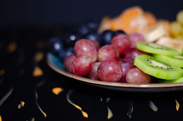 Fruits, grapes, tangerines, bananas, kiwi in a blue porcelain plate. black wooden floor, fruit plate