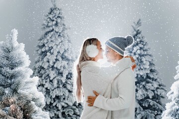 Falling snow and kisses. Happy young couple close-up hugs and kiss near christmas trees at the eve of new year celebration in winter day. Smiley man and woman weared white pullovers love each other