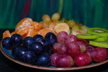 Fruits, grapes, tangerines, bananas, kiwi in a blue porcelain plate. black wooden floor, fruit plate