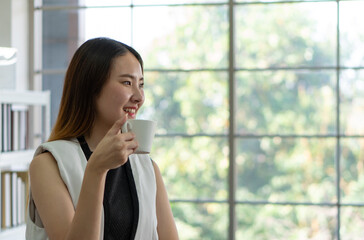 young asian businesswoman taking break and drinking coffee near window in the office. business concept