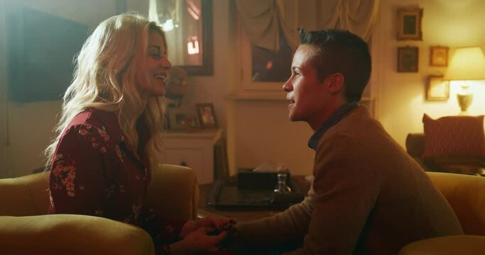 Cinematic shot of young carefree happy smiling homosexual female gay and transgender man couple in love is having fun to speak to each other and kissing while sitting on a sofa in living room at home.