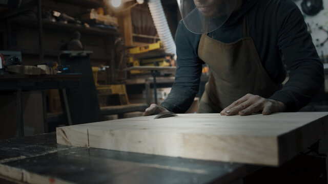 A carpenter wearing an apron and a mask is sawing a tree on a circular saw in a workshop