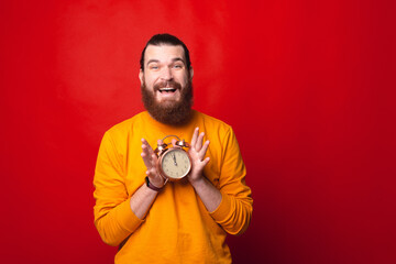 Cheerful bearded man is holding a clock with both hands looking at the camera .