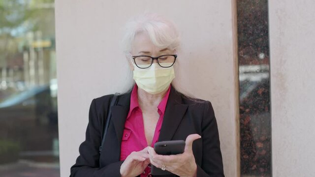 A mature woman wearing a mask due to COVID19 standing in an outdoor urban environment looking at her smartphone switches from reading glasses to sunglasses and then walks past camera.