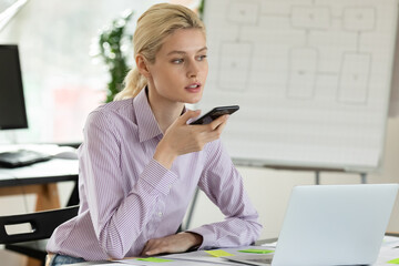 Concentrated young female employee office manager working on computer, sending voicemail, dictating audio message or activating voice assistant in mobile phone application, multitasking concept.