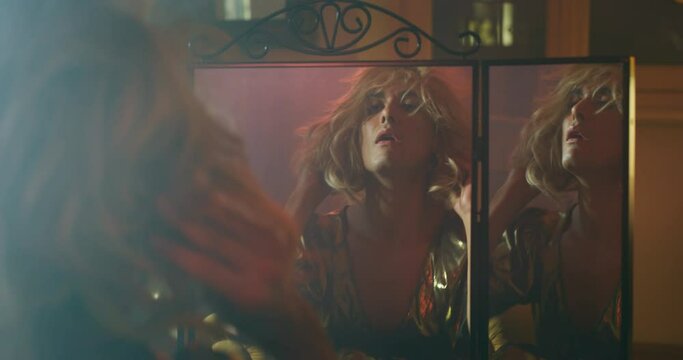 Cinematic shot of an young carefree homosexual male gay is putting on wig and in a mirror while preparing in vanity room. Concept of gender expression, identity and diversity.
