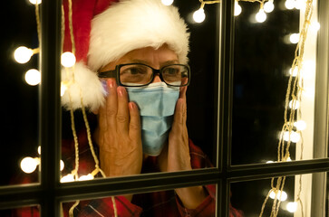 A sad elderly woman behind the window in the night with a Santa hat wears a medical mask due to the coronavirus. Self isolation away from everyone to avoid contagion