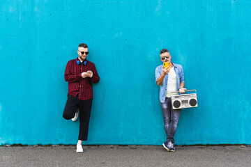 Cool men looking at mobile phone lean against blue wall - Couple of influencer guys listening music with vintage boombox stereo outdoor in the street - Concept of tech, social network and music trends