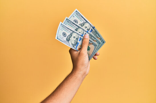 Hand of hispanic man holding 100 dollars banknotes over isolated yellow background.