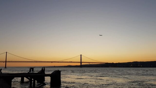 Pier at sunset with silhouette of young people, suspension bridge on the background and airplane flying