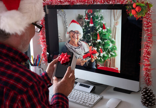 Two smiling senior people with santa hats in video call with computer opening together the Christmas gifts. Old active retirees addicted with new technologies. Lockdown due to coronavirus covid-19