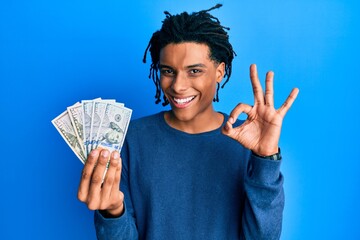 Young african american man holding dollars doing ok sign with fingers, smiling friendly gesturing...