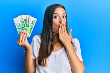 Young hispanic woman holding israel shekels covering mouth with hand, shocked and afraid for...