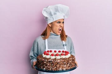 Hispanic young chef woman holding chocolate cake angry and mad screaming frustrated and furious, shouting with anger. rage and aggressive concept.