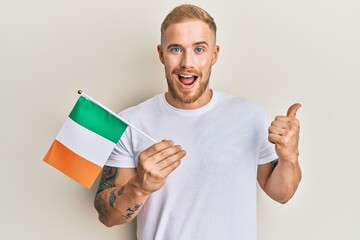 Young caucasian man holding irish flag pointing thumb up to the side smiling happy with open mouth