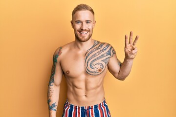 Young caucasian man wearing swimwear shirtless showing and pointing up with fingers number three while smiling confident and happy.